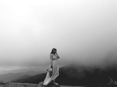 grayscale photo of standing woman wearing wedding dress holding flower bouquet across hills thumbnail