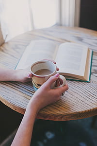 woman holding half-filled coffee cup and book on table thumbnail