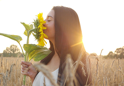 woman wearing white top holding yellow sunflower while standing on brown field during daytime thumbnail