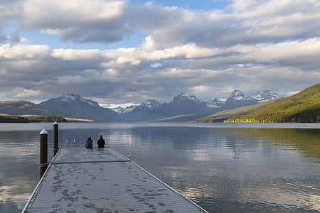 two person sitting on black wooden dock while looking at mountain with body of water scenery during daytime thumbnail