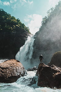 person standing on large brown rock along waterfalls in mountain thumbnail