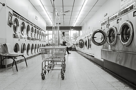grayscale photo of woman carrying plastic laundry basket towards front load commercial washer thumbnail
