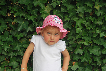 girl wearing pink Hello Kitty bucket hat and white cap-sleeved blouse standing behind green leaves thumbnail