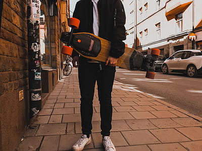 man wearing black jacket and black pants carrying brown longboard thumbnail