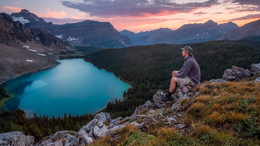 man sitting on rocky hillside watching lake near mountain during daytime thumbnail