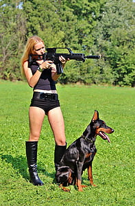 woman holding a rifle with black and tan Doberman pinscher during daytime thumbnail