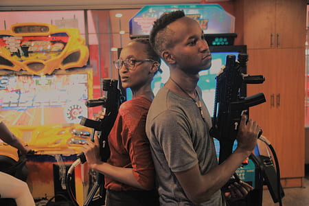 man and woman standing back to back while holding two black game arcade rifle controllers thumbnail