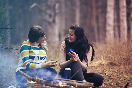 selective focus photograph of two woman sitting on field while holding drinking cups thumbnail