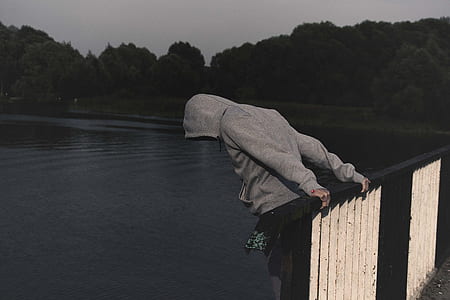 person wearing grey pullover hoodie holding on brown and white metal railings overlooking body of water thumbnail