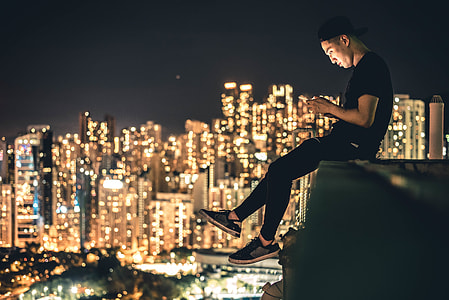 photo of man in black cap, T-shirt, pants, and low top sneakers sitting on edge with high rise buildings with lights during night time thumbnail