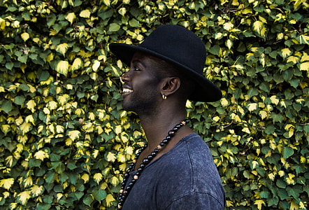 smiling man wearing beaded necklace and black hat standing near yellow and green leaf plant thumbnail