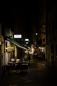 man standing on street with tables and chairs between buildings during night time thumbnail