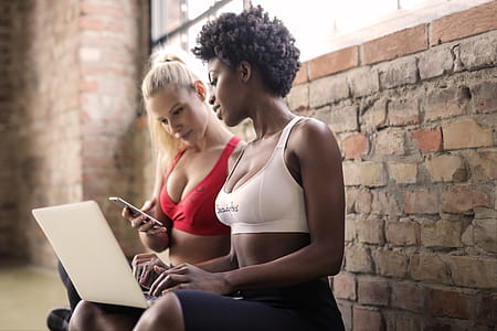 Two multiethnic women wearing sports bra looking at mobilephone and laptop in the gym against a brick wall thumbnail