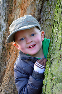 smiling boy wearing gray cap and jacket peeking beside tree trunk with moss thumbnail