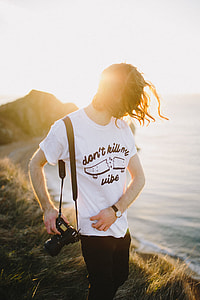 shallow focus photography of man holding DSLR camera on mountain with view of large body of water during daytime thumbnail