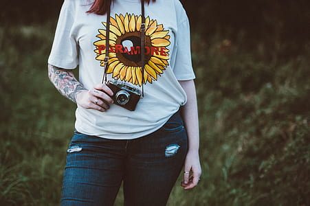 woman wearing white and yellow Paramore crew neck t-shirt holding black MILC camera while standing on grass land at daytime thumbnail