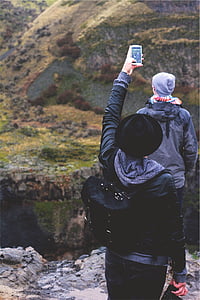 person raising hand while holding smartphone taking photo of man standing near cliff during daytime thumbnail