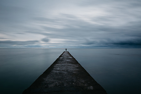 person stand on ocean dock near body of water under cloudy sky thumbnail