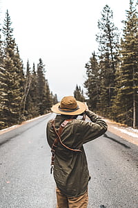 man standing on road taking picture between trees during daytime thumbnail