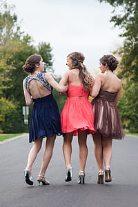three women wearing cocktail dresses talking while walking on grey concrete road between green leaf trees during daytime thumbnail