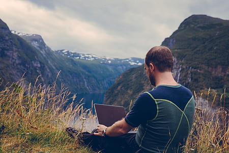 man holding laptop sitting on grass field facing valley with river thumbnail