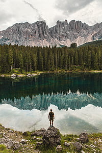 man standing on boulder facing lake and pine trees at daytime thumbnail