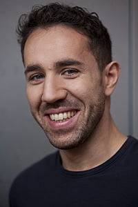 smiling man in black crew neck shirt beside a white wall thumbnail