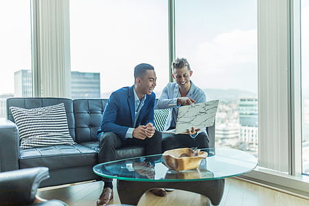 two men laughing sitting on sofa facing laptop near glass building window thumbnail