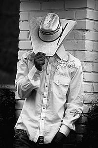 grayscale photography of man wearing floral dress shirt and cowboy hat leaning on bricked wall thumbnail