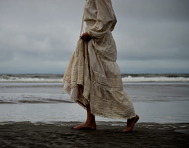 woman wearing beige pleated dress walking on seashore under thick gray clouds photo thumbnail