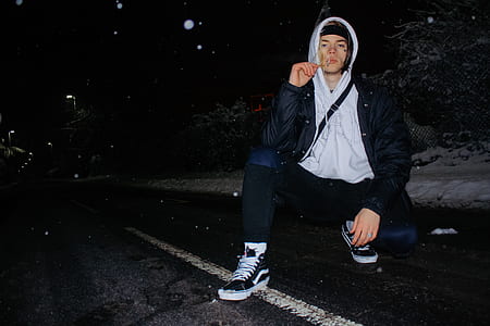 man wearing black and gray button-up hoodie and blue denim jeans sitting on asphalt road during nighttime thumbnail