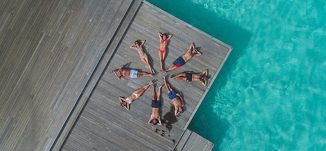 aerial photography of group of people making circle formation while lying on dock beside body of water thumbnail