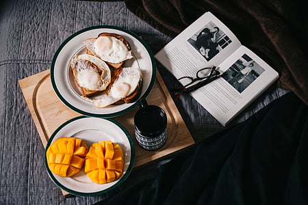 sliced mangoes and sandwich with eggs on brown wooden board beside open book and eyeglasses thumbnail
