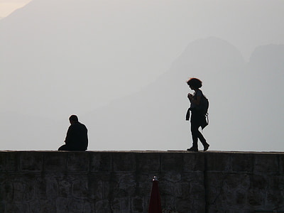 silhouette of person walking towards of man sitting on cliff thumbnail