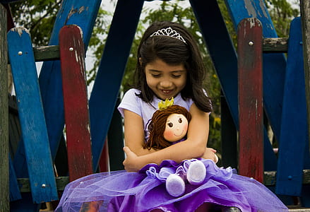 girl in purple t-shirt dress holding rag doll during daytime thumbnail