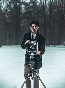 selective focus photography of man wearing black coat using black and grey camera with tripod near tall trees during daytime thumbnail