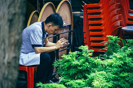 man sitting on red stool while using smartphone beside stacked chairs and folded tables thumbnail