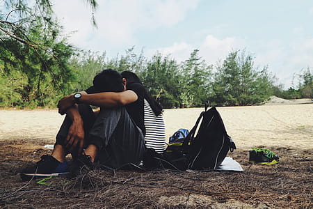 Man in Black and White Striped Shirt With Black Pants and Pair of Black Shoes Sitting on Black Textile Beside Backpack Near Tree thumbnail