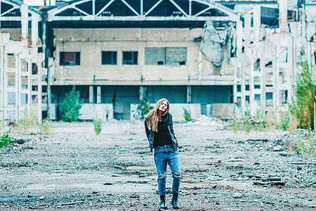 woman in black jacket and blue jeans standing on brown dirt with hands in back pockets thumbnail