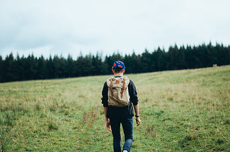 man in black sweater, blue jeans and brown backpack walking on green grass beside forest thumbnail