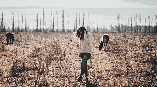 woman raising her left foot while standing between dried grasses with horses under clear blue sky thumbnail
