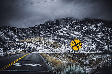 yellow and black road sign beside gray concrete road with view of snow-capped mountain photo thumbnail