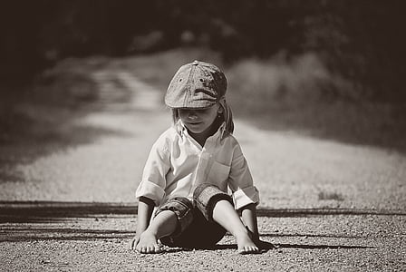 greyscale photography of boy wearing dress shirt and newsboy cap sitting on ground at daytime thumbnail
