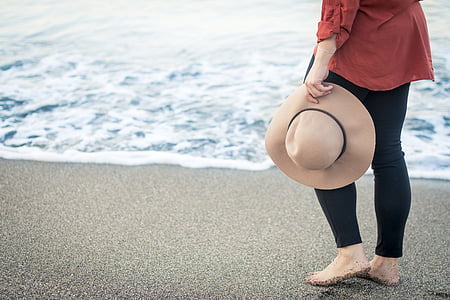 woman holding brown sunhat standing on gray sand beside sea during daytime thumbnail