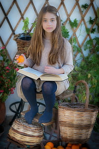 girl in gray knit sweater holding book and orange fruit sitting on chair beside brown wicker basket thumbnail