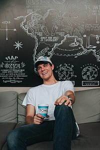 man sitting on gray couch wearing white crew-neck t-shirt holding TOMS cup over painted wall thumbnail
