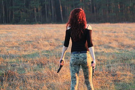 woman wearing black cold-shoulder quarter-sleeved shirt with green and black camouflage pants holding two black semi-automatic pistols standing on brown and green grass field thumbnail