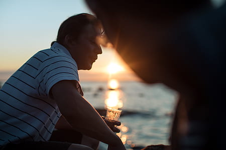 man holding drinking glass filled with liquid near body of water at daytime thumbnail