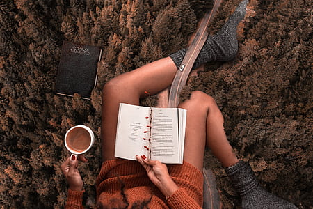 woman wearing orange sweater and grey socks reading a book with white mug thumbnail