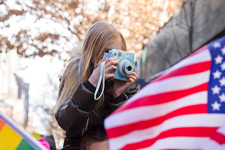 woman wearing bubble jacket using camera near U.S. flag during daytime thumbnail